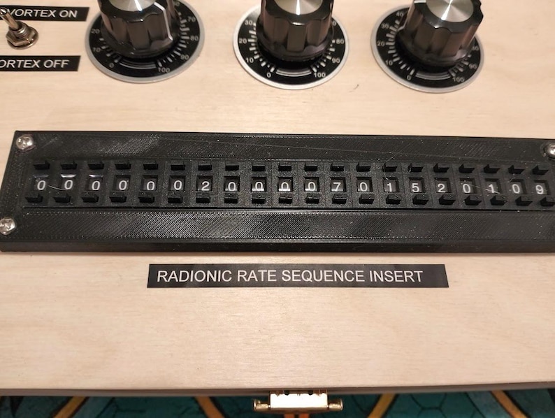 May include: Close-up of a wooden panel with black dials, switches, and a black digital display. The display shows a series of numbers and letters. The text "RADIONIC RATE SEQUENCE INSERT" is printed below the display.