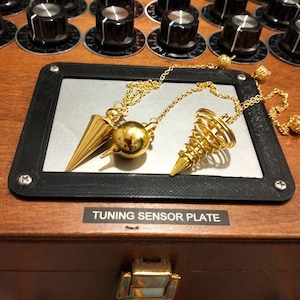 May include: A wooden box with a Tuning Sensor Plate label, featuring a silver panel with two gold-coloured pendulums. The box has several black knobs on top. The pendulums have a cone and spiral design.