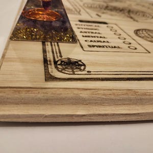 May include: A wooden board with etched designs and text, including a pyramid-shaped object with purple and gold accents. The board features geometric patterns and the words "PHYSICAL", "ASTRAL", "MENTAL", "CAUSAL", and "SPIRITUAL".