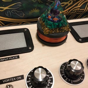 May include: A wooden panel with black framed plates labeled "INPUT PLATE" and "OUT PLATE". A pyramid-shaped object sits atop a coil of copper wire. Knobs with numerical markings and the text "VORTEX ON" are also visible.