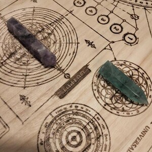 May include: Two crystal wands, one purple and one green, rest on a wooden surface etched with a circular diagram and the word "OUTPUT". The wands are pointed and appear to be made of polished stone.