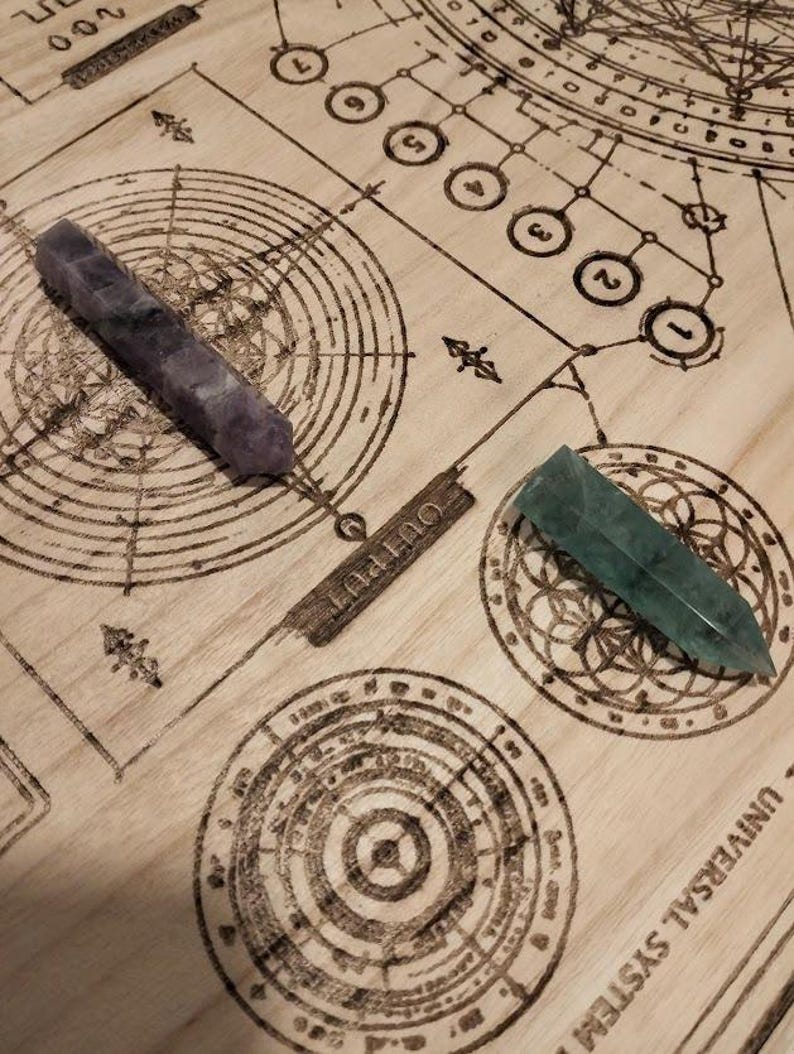 May include: Two crystal wands, one purple and one green, rest on a wooden surface etched with a circular diagram and the word "OUTPUT". The wands are pointed and appear to be made of polished stone.