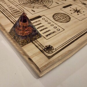May include: A wooden board with etched diagrams and text, including a scale and geometric shapes. A pyramid-shaped object with copper wire and purple and gold accents sits on the corner of the board. The board has a light wood grain.