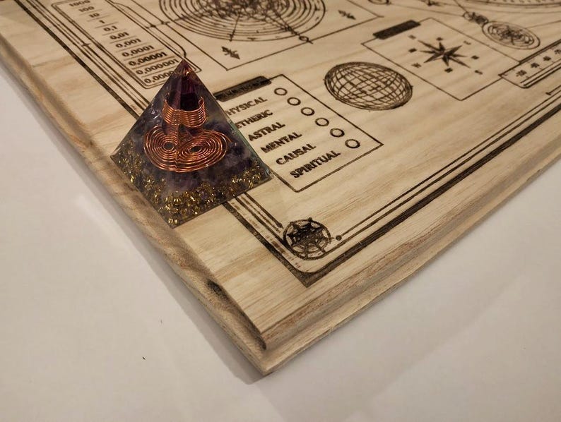 May include: A wooden board with etched diagrams and text, including a scale and geometric shapes. A pyramid-shaped object with copper wire and purple and gold accents sits on the corner of the board. The board has a light wood grain.