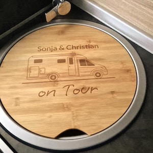 Può includere: Un tagliere rotondo in legno con bordo argentato, con un disegno di camper inciso e le parole "Sonja & Christian" e "on Tour". Il tagliere è inserito in un piano di lavoro.