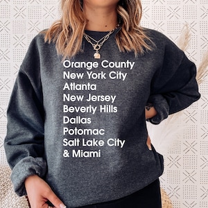 May include: A dark gray sweatshirt with white text listing various cities and locations, including Orange County, New York City, Atlanta, New Jersey, Beverly Hills, Dallas, Potomac, Salt Lake City, and Miami.