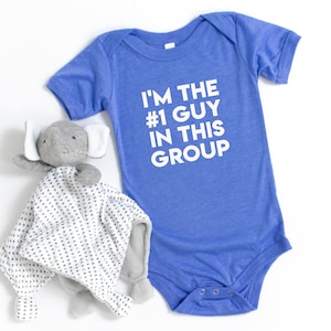 May include: A blue baby onesie with the text "I'm the #1 guy in this group" in white. A gray elephant stuffed animal with a white and gray patterned blanket is in the foreground.
