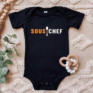 May include: Black baby bodysuit with the words "SOUS CHEF" in orange and white, and a spoon graphic. The bodysuit is displayed on a beige textured surface, with wooden baby toys nearby.