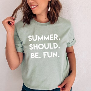 May include: A pale green t-shirt featuring the white text "SUMMER. SHOULD. BE. FUN." in a bold, sans-serif font. The shirt has short sleeves and a casual style. The model is wearing the shirt.