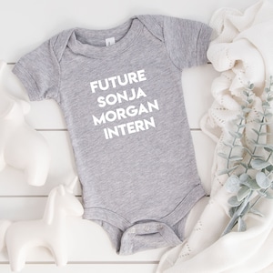 May include: A gray baby onesie with the text "FUTURE SONJA MORGAN INTERN" printed in white.