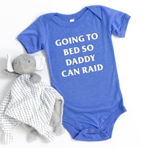 Könnte beinhalten: Ein blauer Baby-Strampler mit dem Text "Going to bed so daddy can raid" in Weiß. Ein graues Stofftier-Elefant mit einer weiß-grauen gemusterten Decke befindet sich im Vordergrund.