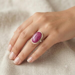 May include: A silver ring featuring a large, smooth, oval-shaped magenta gemstone set in a simple metal bezel. The stone has a polished surface with subtle natural variations in color and texture, securely mounted on a plain, thin silver band.