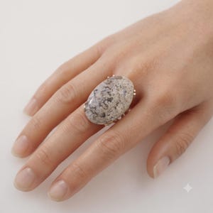 Puede incluir: Un anillo con una piedra grande de forma ovalada que presenta un patrón moteado y jaspeado en tonos gris, beige y blanco. La piedra está montada en una banda metálica con garras visibles que aseguran la gema, mostrando un aspecto natural.
