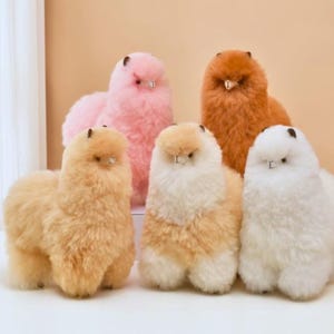 May include: A collection of five fluffy alpaca plush toys in various colors: pink, orange, white, and two shades of tan. The alpaca toys have small, dark eyes and ears, and are standing on a white surface.