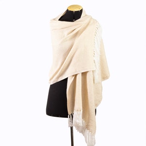 May include: A cream-colored shawl with a geometric pattern and white fringe. The shawl is draped over a black mannequin.