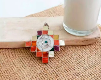 May include: A silver pendant with a colorful mosaic design in the shape of a cross. The center of the cross features a white shell with a turquoise eye.