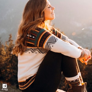 May include: A woman wearing a white sweater with a colorful geometric pattern, black pants, and patterned socks. She is sitting on a rock with a mountain background.