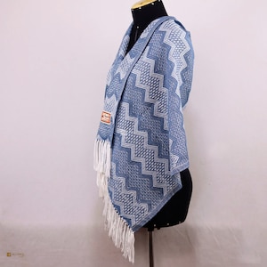 Puede incluir: Un poncho de alpaca azul y blanco con un patrón de zigzag y flecos blancos. El poncho tiene una capucha y se muestra en un maniquí.