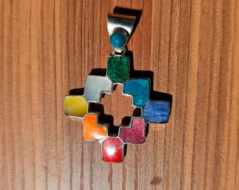 May include: A silver pendant with a colorful, geometric design. The pendant is made of silver and features a variety of gemstones in shades of red, orange, yellow, green, blue, and purple.