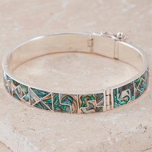 Puede incluir: Un brazalete de plata con un diseño geométrico hecho de concha de abulón iridiscente. El brazalete tiene bisagras y un cierre.