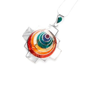 Puede incluir: Un collar de plata con un colgante circular de varios colores y capas. El colgante está hecho de varias piedras y está engastado en una cruz de plata.