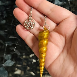 May include: A gold-colored spiral shell pendant attached to a silver-toned chain, featuring a small circular silver-toned charm with a five-pointed star pentagram design. The pendant hangs from a metal jump ring connected to the chain links.