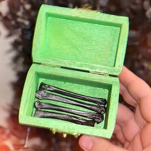 May include: A small, open, green wooden box with a hinged lid, containing several dark, bone-shaped objects. The box is held in a hand, and the background is blurred with dark green and brown tones.