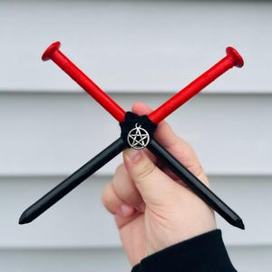May include: Two crossed metal spikes, one painted bright red and the other painted glossy black, are bound together at the center with black cord. A small, circular silver-toned metal charm featuring a five-pointed star pentagram hangs from the center.