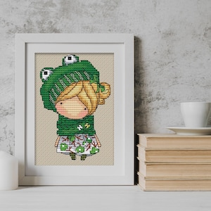 May include: Cross-stitch pattern of a girl wearing a green frog hat and a green and white dress. The girl has blonde hair and is smiling.