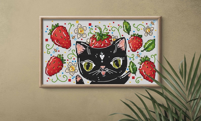 May include: Cross-stitch art featuring a black cat with a strawberry on its head, surrounded by red strawberries, white flowers, and green leaves. The artwork is framed in a light brown wooden frame.