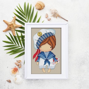 May include: A framed cross-stitch of a sailor doll with red hair, wearing a blue and white sailor outfit and hat. The doll is holding a small boat. The frame is white, and the background includes seashells, a starfish, and palm leaves.