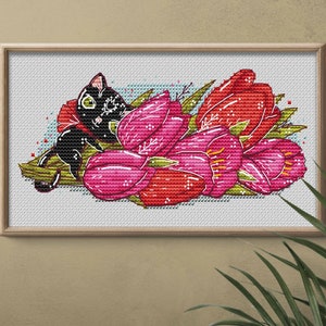 May include: Cross-stitch art featuring a black cat resting among vibrant red and pink tulips. The framed artwork has a light wood frame and is set against a neutral background. The cat has green eyes and is decorated with white patterns.