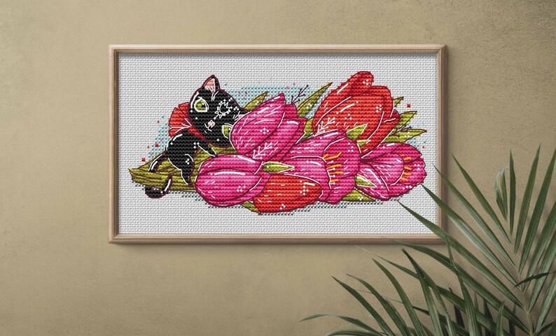 May include: Cross-stitch art featuring a black cat resting among vibrant red and pink tulips. The framed artwork has a light wood frame and is set against a neutral background. The cat has green eyes and is decorated with white patterns.