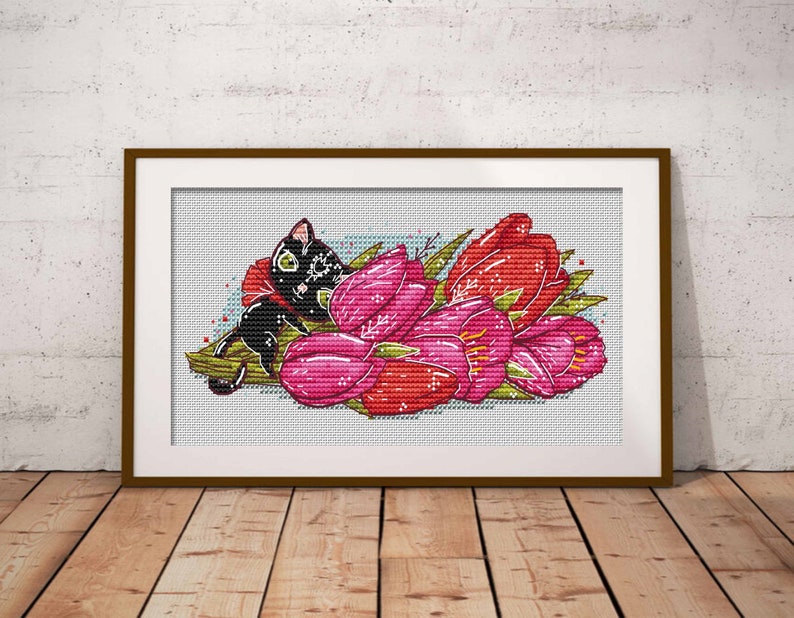 May include: Cross-stitch art of a black cat nestled among pink and red tulips. The cat has green eyes and is wearing a red bow. The artwork is framed in a brown wooden frame and set against a white background.
