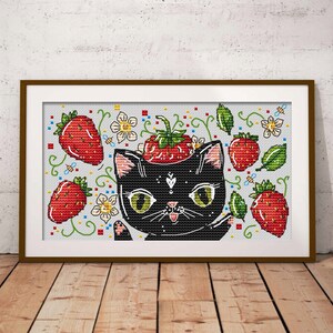 May include: Cross-stitch art featuring a black cat with green eyes and a strawberry on its head. The design includes red strawberries, green leaves, white flowers, and bees. The artwork is framed in a brown wooden frame and set against a white brick wall.