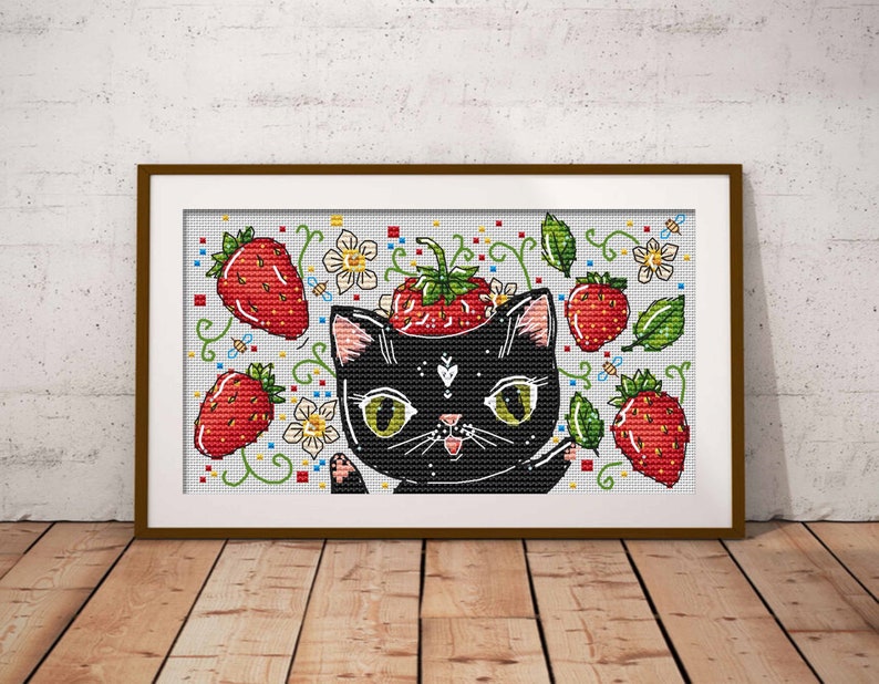 May include: Cross-stitch art featuring a black cat with green eyes and a strawberry on its head. The design includes red strawberries, green leaves, white flowers, and bees. The artwork is framed in a brown wooden frame and set against a white brick wall.