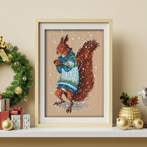 May include: A framed cross-stitch design of a squirrel wearing a blue and white sweater with a brown tail. The squirrel is standing on a beige background with white snowflakes.