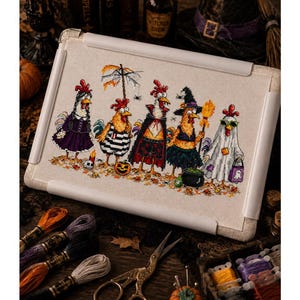 May include: A cross-stitch embroidery project on a rectangular frame featuring five chickens dressed in Halloween costumes, including a vampire, a witch, and a ghost, standing on a bed of autumn leaves with a small pumpkin and a black cauldron.