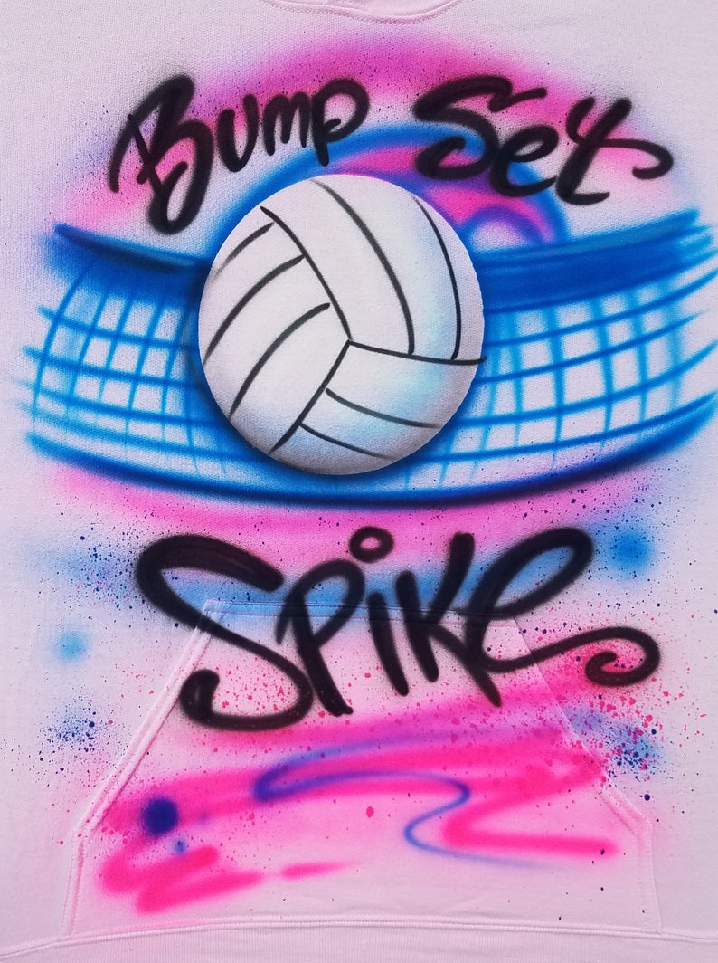 May include: A light pink hoodie with a volleyball-themed design. The design features a white volleyball, a blue net, and the words "Bump Set Spike" in black script, with pink and blue spray paint accents.