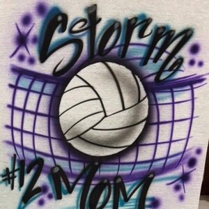 May include: A light gray t-shirt featuring an airbrushed volleyball design. The design includes a volleyball, net, and the word "Storm" in black and blue script. Below the volleyball is the text "#12 Mom".