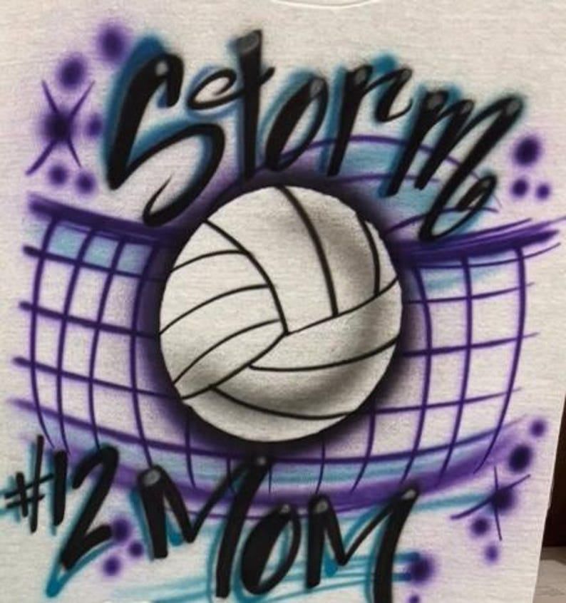 May include: A light gray t-shirt featuring an airbrushed volleyball design. The design includes a volleyball, net, and the word "Storm" in black and blue script. Below the volleyball is the text "#12 Mom".