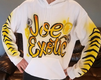 May include: White hoodie with a yellow and black tiger stripe design on the sleeves. The front of the hoodie has a yellow airbrushed design that says "Joe Exotic".