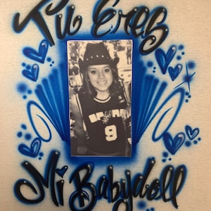 May include: A white t-shirt with a blue and black airbrushed design. The design features a photo of a person wearing a cowboy hat and a basketball jersey, surrounded by hearts and the words "Tu Eres Mi Babydoll".