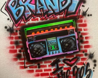 May include: A white t-shirt with a graffiti design featuring the name "BRANDI" in blue, pink, and green. A boombox with a green, orange, and pink design is below the name. The words "I love the 90s" are below the boombox.