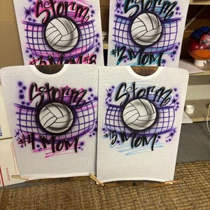 May include: Four white t-shirts with airbrushed volleyball designs. Each shirt features a volleyball, the word "Storm", and the text "#2 Mom", "#3 Mom", "#4 Mom", and "#5 Mom". The designs include purple, pink, and blue accents.