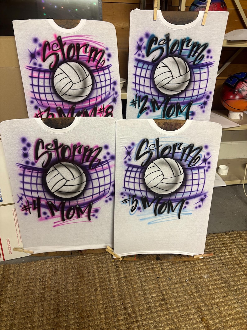 May include: Four white t-shirts with airbrushed volleyball designs. Each shirt features a volleyball, the word "Storm", and the text "#2 Mom", "#3 Mom", "#4 Mom", and "#5 Mom". The designs include purple, pink, and blue accents.