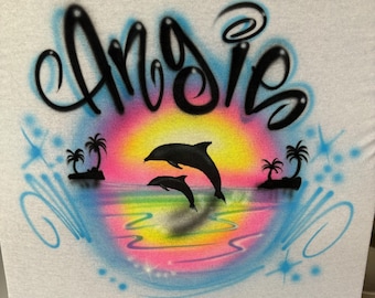 May include: A white t-shirt with a custom airbrushed design featuring the name "Angie" in black script, a sunset with pink, orange, and yellow hues, and two dolphins jumping out of the water.