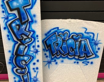 May include: A gray hoodie with a blue airbrushed design of the name "TRICIA" in a graffiti style. The hoodie has a pocket on the front and a hood.