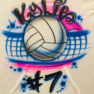 May include: White hoodie with an airbrushed volleyball design. The design includes the name "Kylie" in black script above a volleyball, a blue net, and the number "7" below. Pink and blue accents complete the design.