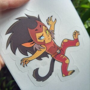 May include: A cartoon sticker of a red and yellow lion-like character with a black mane, wearing a red and brown outfit. The character is in a dynamic pose with one leg raised and claws extended.